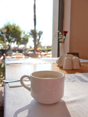 A white ceramic coffee mug sits on a sunlit cafe table by a large window. The outdoor scene is softly blurred, creating a calm, cozy mood perfect for breakfast and relaxation.