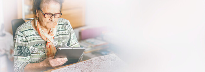 An elderly woman sits indoors and focuses on using a tablet, illuminated by warm natural light.