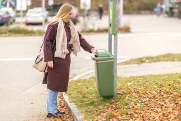 Woman disposing waste in outdoor public bin during autumn, dressed in coat and scarf, practicing...