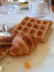 A delicious breakfast plate featuring a flaky croissant, waffle, and soft pancakes with a light glaze and orange garnish, set on a white table for a warm, cozy family morning.