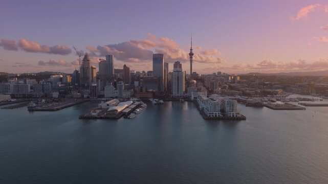 Aerial view of Auckland, New Zealand, at sunset. The city skyline features the Sky Tower, modern buildings, and a harbor with boats. The photo captures the beauty of the city.