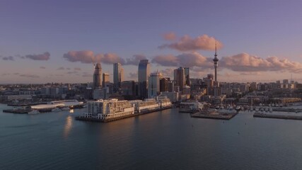 Auckland, New Zealand skyline at dusk. The city's skyscrapers and the Sky Tower are visible against a backdrop of pink and purple clouds. The harbor is calm, reflecting the city lights.