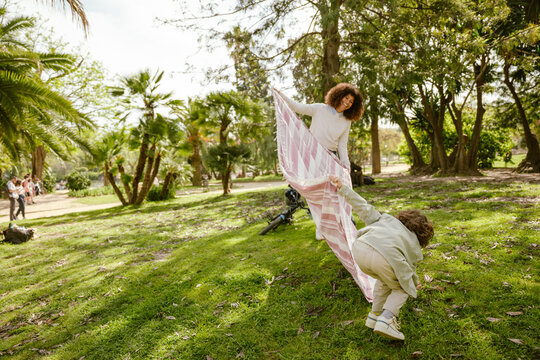 A mother laughs and looks at her son who is standing in front of her, bent over, while they hold a blanket