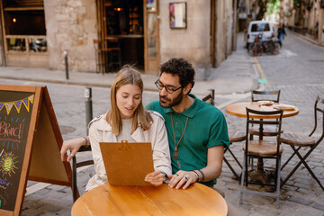 A man and a woman are sitting at a table and reading a menu that she is holding while talking