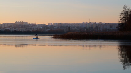 Naklejka premium A person paddles a board on water. A cityscape sits distant. Reeds line the shore. The sky glows orange. Reflections appear on the surface. Trees are visible. It is a ser