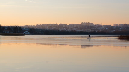 Naklejka premium A person standup paddleboarding on a calm body of water. Buildings are visible on the distant shore. The sky exhibits a warm, golden hue. Reflections appear on the water'