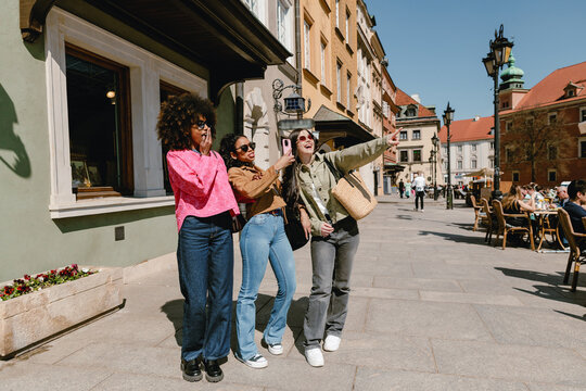 A group of three female friends stand and look in the direction one of them is pointing while one of them covers her mouth with her hand and the other holds a phone while they talk and laugh