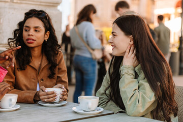 Two female friends sitting at a table and listening while one smiles and the other talks and holds a cup