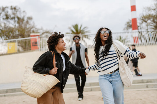 Two female friends are running and holding hands while laughing and their male friend is walking behind them