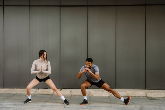 A female athlete talks to a male athlete next to her as they squat and stretch their legs while holding their hands together in front of them - Powered by Adobe