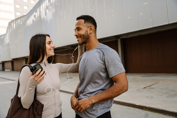 Female and male athlete listening to earphones and smiling while standing next to each other and she holding a phone