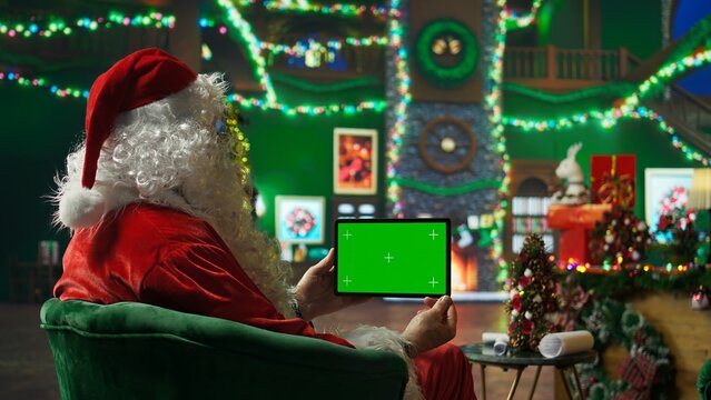 Person dressed as Santa Claus reading letters from kids on green screen tablet. Senior man portraying Christmas character answering children using mockup device, camera A