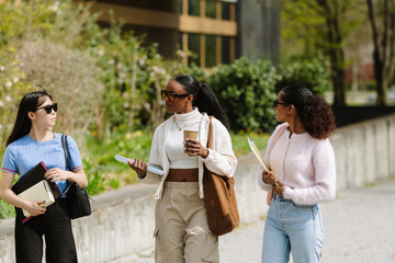 A female student holds a cup and a notebook while talking and listening to two female students walking next to her and holding folders