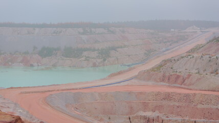 Quarry landscape shows a turquoise pool. Slopes display varied rock layers. A conveyor belt transports material. Trees line the distant edge. Dust hangs in the hazy air.