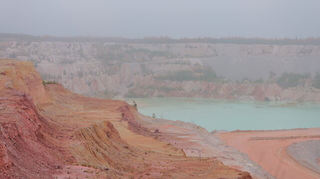 Quarry landscape shows vibrant hues. Mineral deposits create colorful strata. Water body reflects sky. Terrain exhibits erosion. Vegetation appears sparse. Aerial view hi