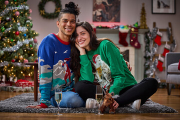 Happy couple wearing festive ugly Christmas sweaters poses together in decorated living room with tree
