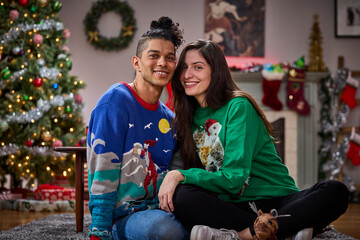 Happy couple wearing festive ugly Christmas sweaters posing together in decorated holiday living room