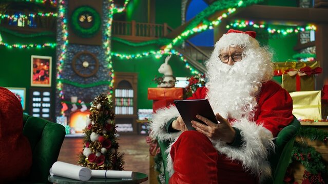 Elderly man dressed as Santa Claus looking at letter received from kids on tablet. Older person wearing Santa costume reads wish lists written by children for Christmas on digital device, camera A