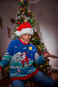 Man wearing Santa hat and festive surfing Christmas sweater poses in front of decorated tree
