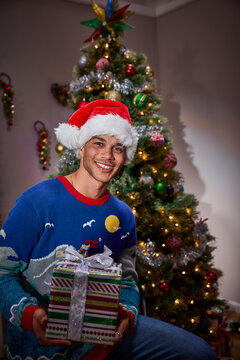 Man wearing Santa hat and festive sweater holding wrapped Christmas gift by decorated tree