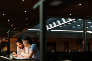 Two female students sitting at a table and smiling while one of them reads a book and the other looks at a laptop