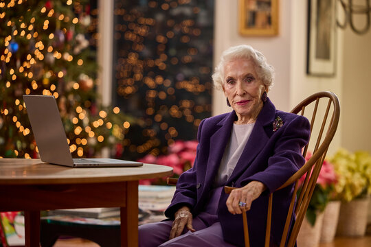 Senior woman in purple suit sitting by laptop with Christmas tree lights in background