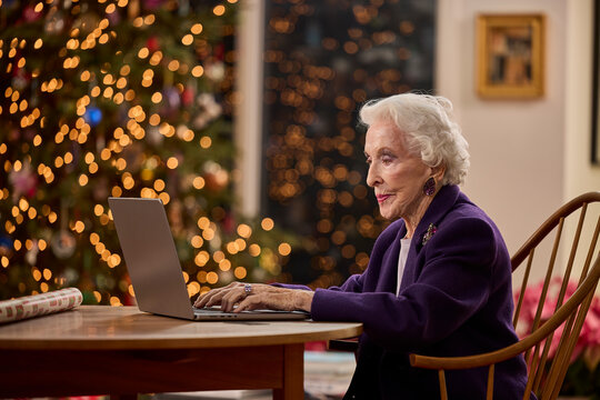 Senior woman using laptop at home during Christmas holiday season with festive tree lights