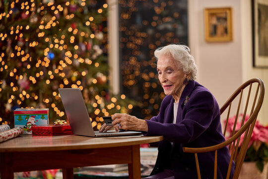 Senior woman using laptop for online holiday shopping beside decorated Christmas tree with twinkling lights