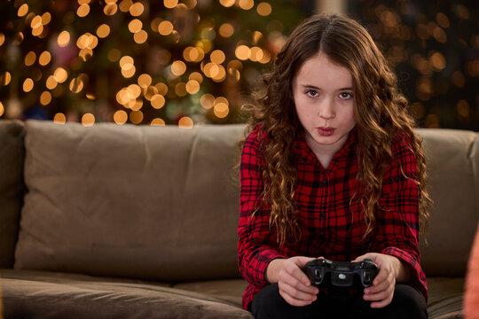 Young girl in red plaid shirt playing video games with Christmas lights in background
