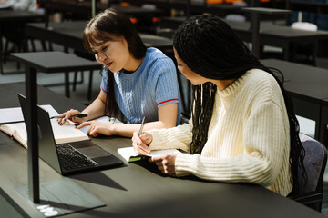Two female students reading a book and smiling while holding pens and sitting at a table