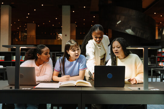 A group of four female students are looking at a laptop, one of them is pointing while the other three are sitting at a table - Powered by Adobe