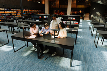 A group of four female students are reading a notebook held by one of them, who is sitting at a table next to two others who are holding a book and another one is standing next to them