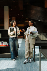 Two female students gesturing and talking as they walk, one holding a laptop and the other a notebook