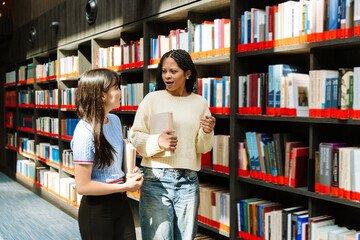 Two female students talking and holding books while walking near bookshelves