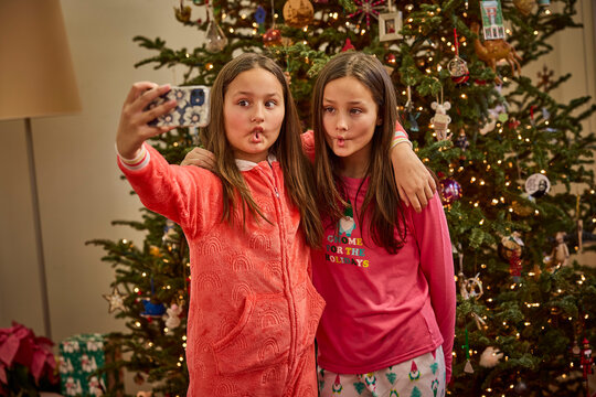 Two girls in pajamas take a selfie while making duck faces in front of Christmas tree