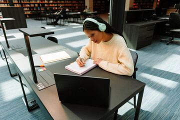 Female student looking at laptop and writing in notebook while sitting at desk and listening to headphones