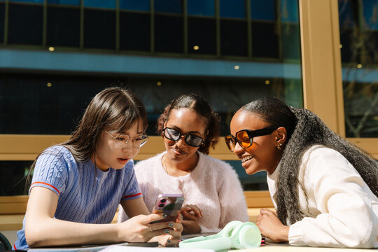 A group of three female students are sitting at a table and looking at a phone that one of them is showing and talking to while two of them are smiling
