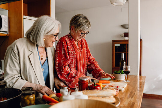 Two female friends laughing and standing at a table while one of them cuts carrots with a knife