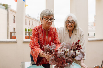 A woman adjusts plants in a vase while standing at a table next to her female friend who is talking while they smile