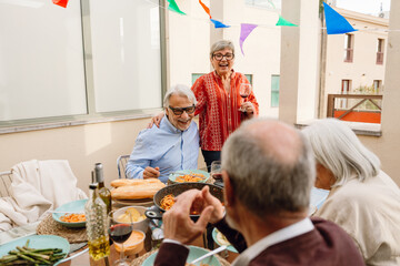 A woman and a man are laughing and hugging while she stands and holds a glass and he sits at a table with two friends