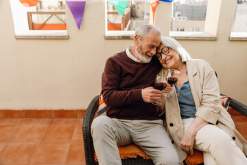 A man and a woman sit on a chair and lean on each other while holding glasses and smiling