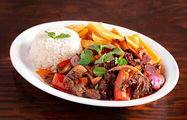 Peruvian beef with roasted vegetables, yuca fries and white rice in a white plate on a wooden table; copy space