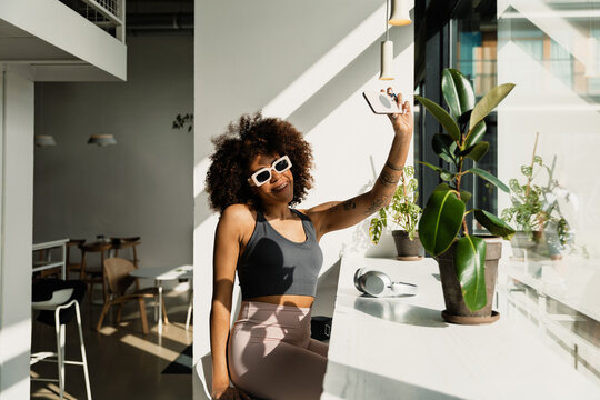 A woman smiles and poses in front of a phone she is holding while sitting on a chair at a table