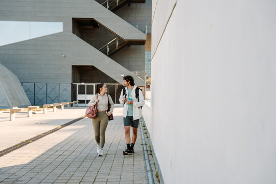 Male and female athletes walking and looking at each other while he holds a bottle and she holds a bag and mat on her shoulder - Powered by Adobe