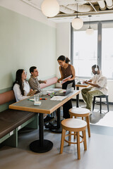A female worker stands and holds a notebook while talking to two workers sitting at a table while they laugh and a male worker sits and types on a laptop next to them
