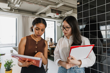 Female worker points to a notebook showing a female worker who is talking and holding a folder while standing next to her