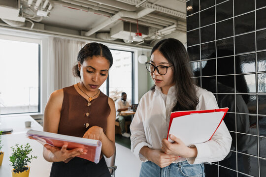 Female worker points to a notebook showing a female worker who is talking and holding a folder while standing next to her