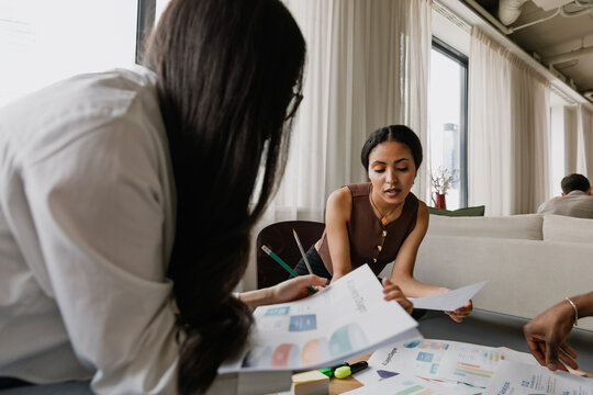 A female employee is talking while sitting at a table next to a female employee and they are holding documents - Powered by Adobe