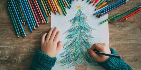 Childs hands draw a Christmas tree with colored pencils on wooden table. Creative holiday activity, cozy atmosphere, handmade traditions.