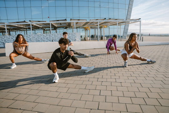 A group of five athletes squat and stretch their legs while holding their hands together in front of them - Powered by Adobe